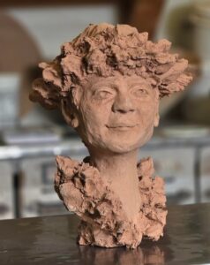 A front-facing view of a clay sculpture of a person's head and neck. The face has a slightly smiling expression, and the hair is sculpted in a thick, textured, cloud-like or popcorn-like mass. The clay is a reddish-brown color and the sculpture sits on a dark surface with a blurred background.
