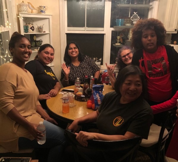 Seven people gather around a wooden dining table in a warmly lit home at night. Several adults smile and wave toward the camera while sitting around the table with drinks and snacks. A child with curly hair stands beside the table wearing a red shirt. Shelves with dishes and decorations and a window with soft lights create a cozy background.