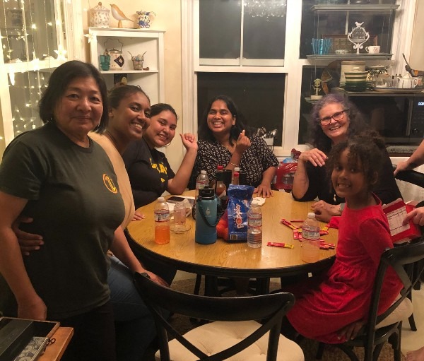 Seven people of different ages gather around a wooden dining table indoors at night, smiling toward the camera. Drinks, snack bags, and water bottles sit on the table. Warm string lights glow near a window in the background, and shelves behind them display dishes and decorative items, creating a cozy, welcoming atmosphere.